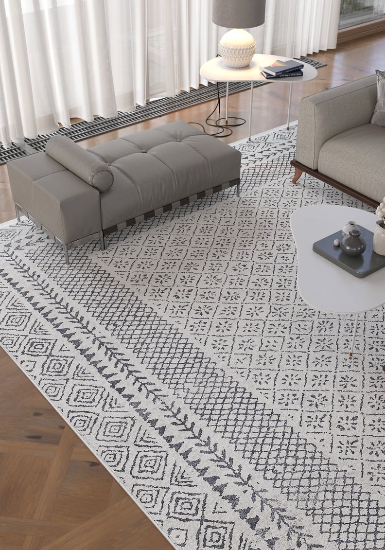 A living room featuring the Classic Contrast - Washable Rug - JR1985 (Custom Size) in gray-anthracite, a gray bench and sofa, a modern white coffee table, a side table with lamp, and sheer curtains that let in natural light.