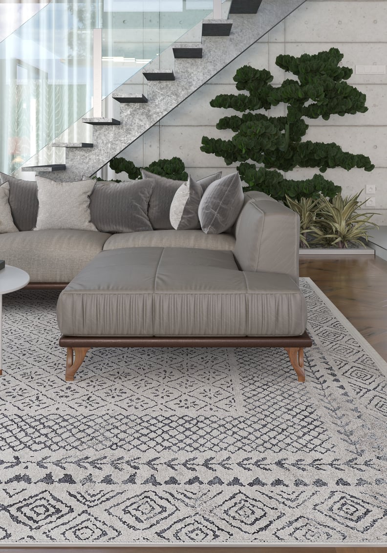 A modern living room features a gray sectional sofa with assorted pillows atop the Classic Contrast - Washable Rug - JR1985 (Custom Size), a potted plant, glass-railed staircase, and large windows letting in natural light.