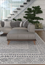 A modern living room features a gray sectional sofa with assorted pillows atop the Classic Contrast - Washable Rug - JR1985 (Custom Size), a potted plant, glass-railed staircase, and large windows letting in natural light.