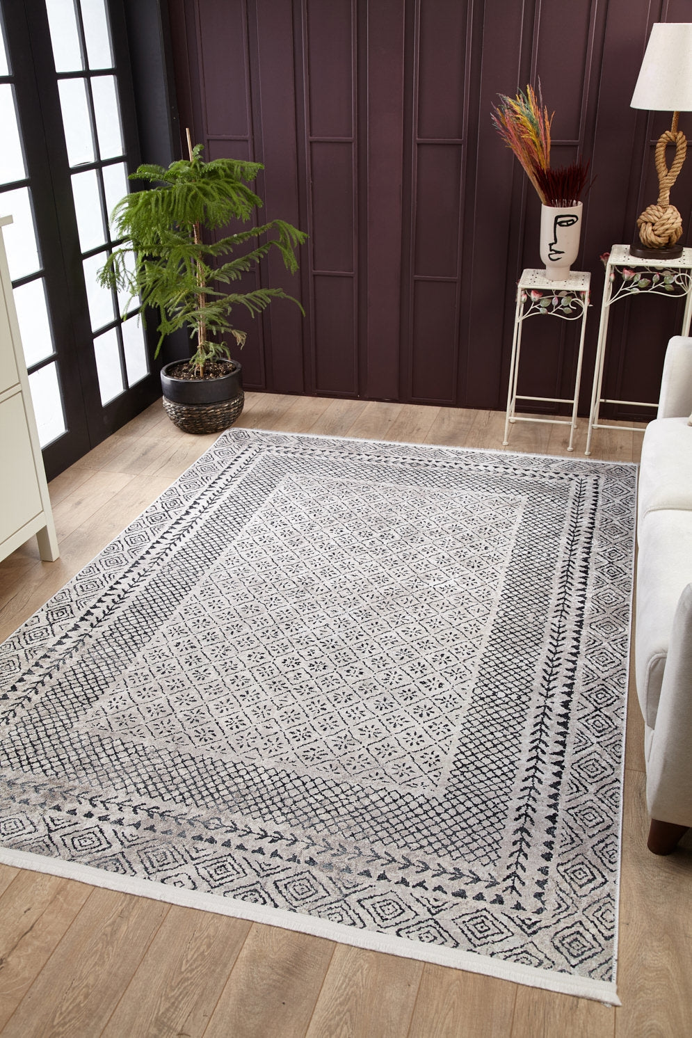 A living room with wooden floors features the Classic Contrast - Washable Rug - JR1985 (Custom Size) in gray-anthracite, a potted plant by large windows, a white sofa, and decorative tables with stylish accents against a dark paneled wall.