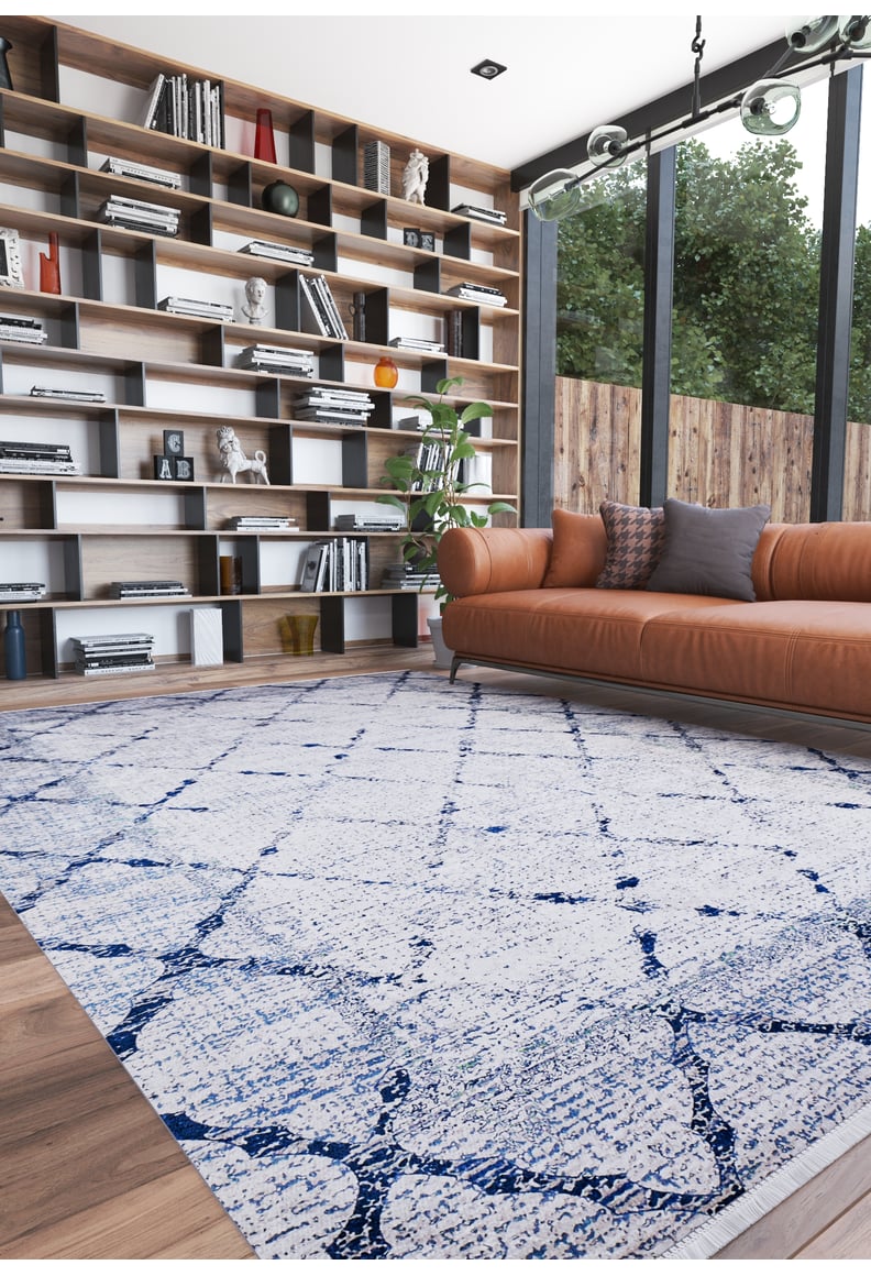A modern living room features a brown leather sofa with cushions, a tall bookshelf, and the Classic Trellis - Washable Rug - JR710 (Custom Size) on a wooden floor. Large windows reveal trees outside.