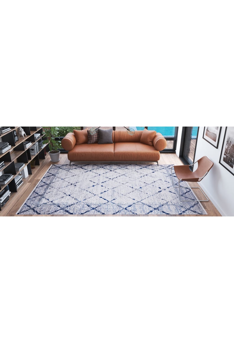 A modern living room with a tan leather sofa, brown accent chair, bookshelves, a potted plant, and the Classic Trellis - Washable Rug - JR710 (Custom Size), featuring blue geometric patterns on a wooden floor.