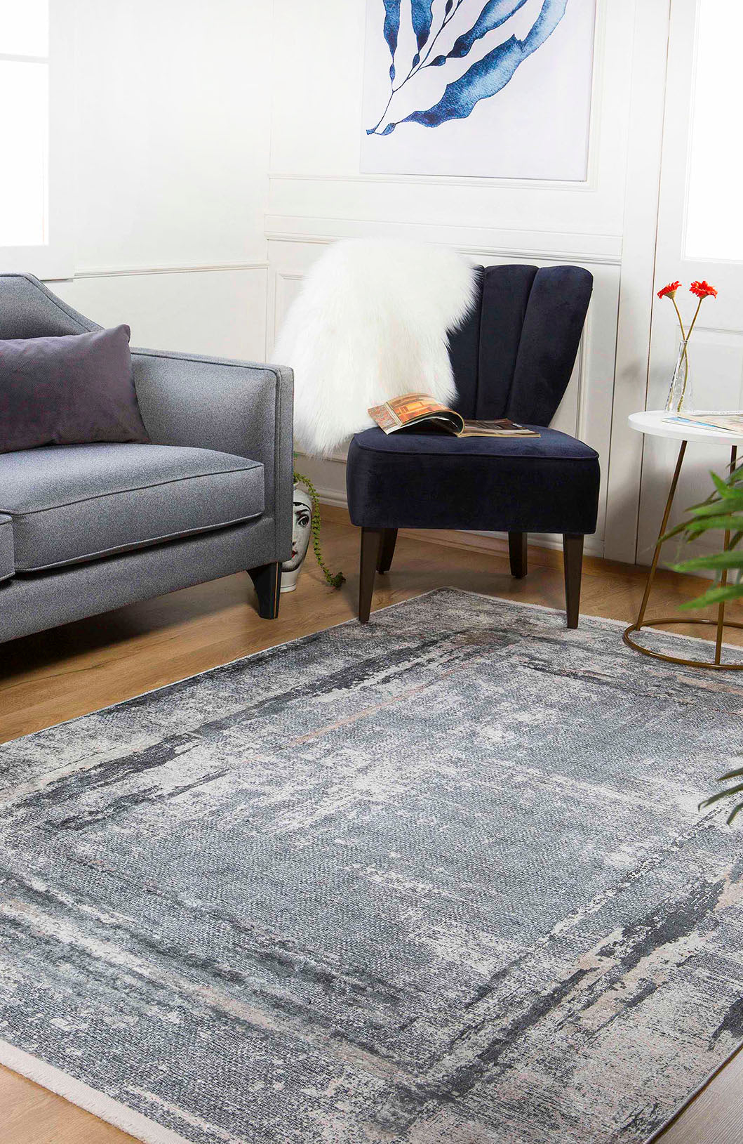 A modern living room features a gray sofa, navy accent chair with a white faux fur throw, the Classic and Bold - Washable Rug - JR1529 (Custom Size), a side table with red flowers, blue leaf art, a plant, and a decorative face sculpture.