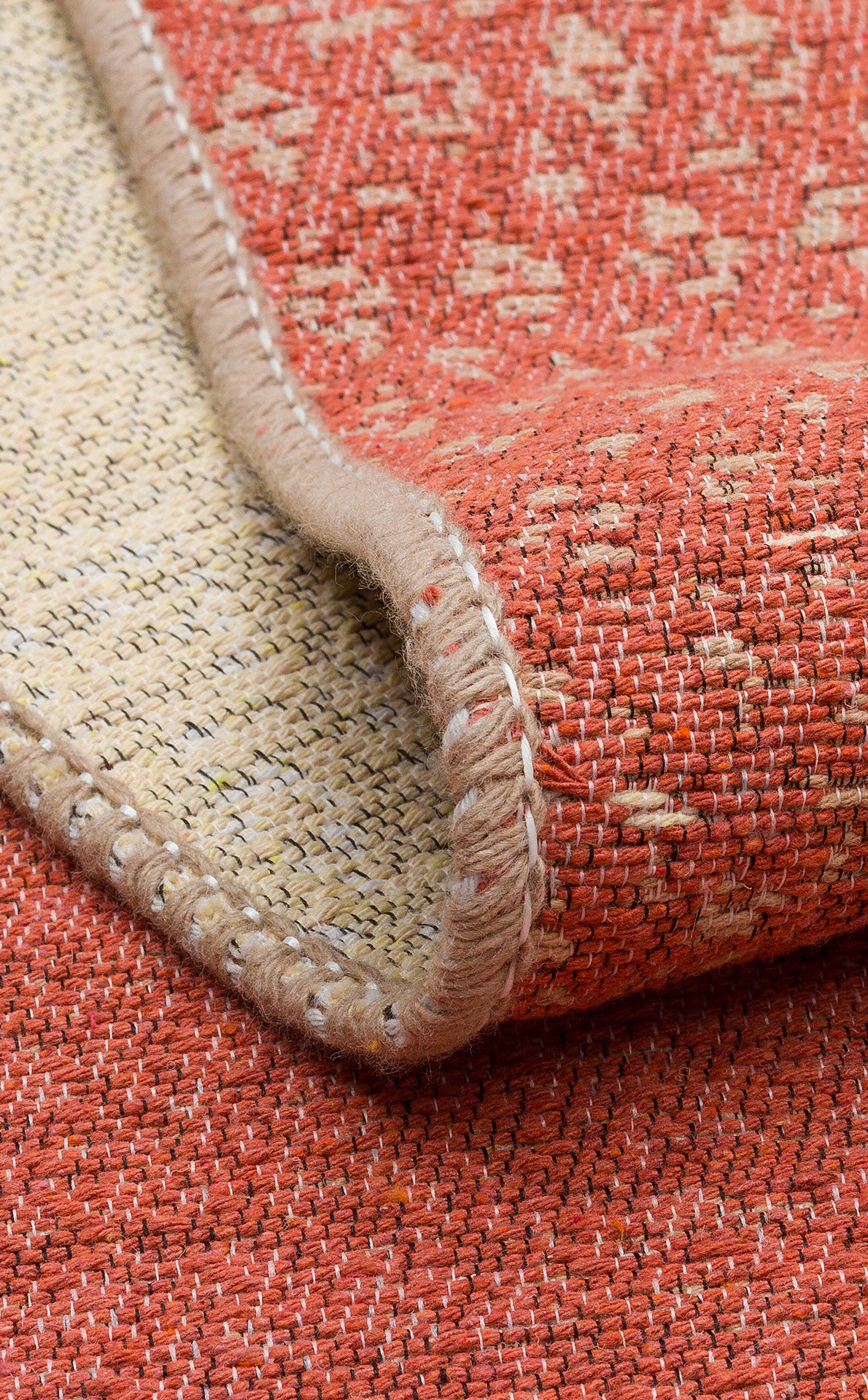 A close-up of the Clay Blend - Washable Double-Sided Kilims - AR37, featuring red and beige tones with a folded corner that highlights its textured surface, detailed stitching, and subtle geometric pattern—ideal for bohemian decor.
