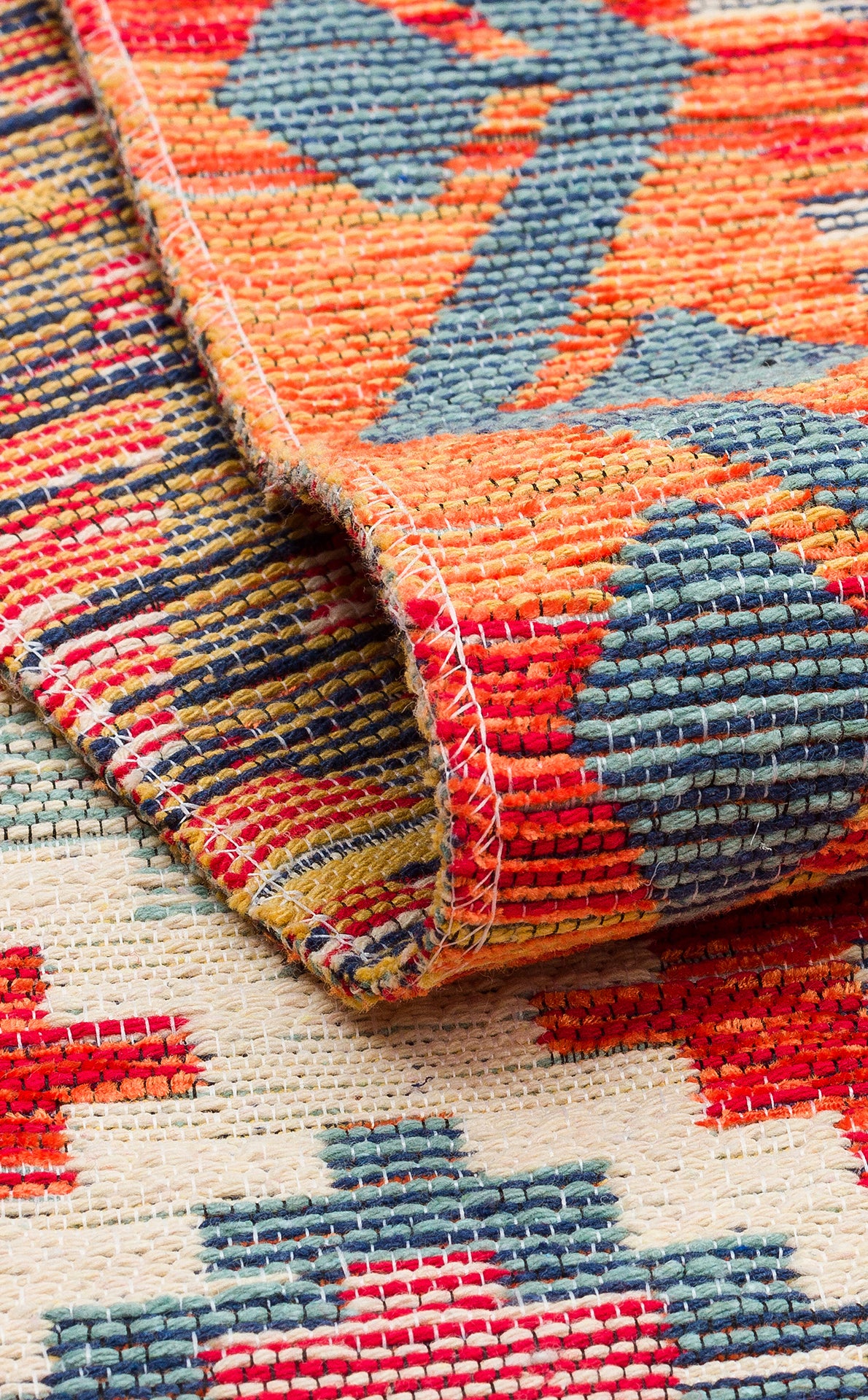 A close-up of the Clay Mark - Washable Reversible Kilim - NMD05 folded at the corner, showcasing geometric patterns in red, orange, blue, and yellow on a cream background with visible texture and stitching.