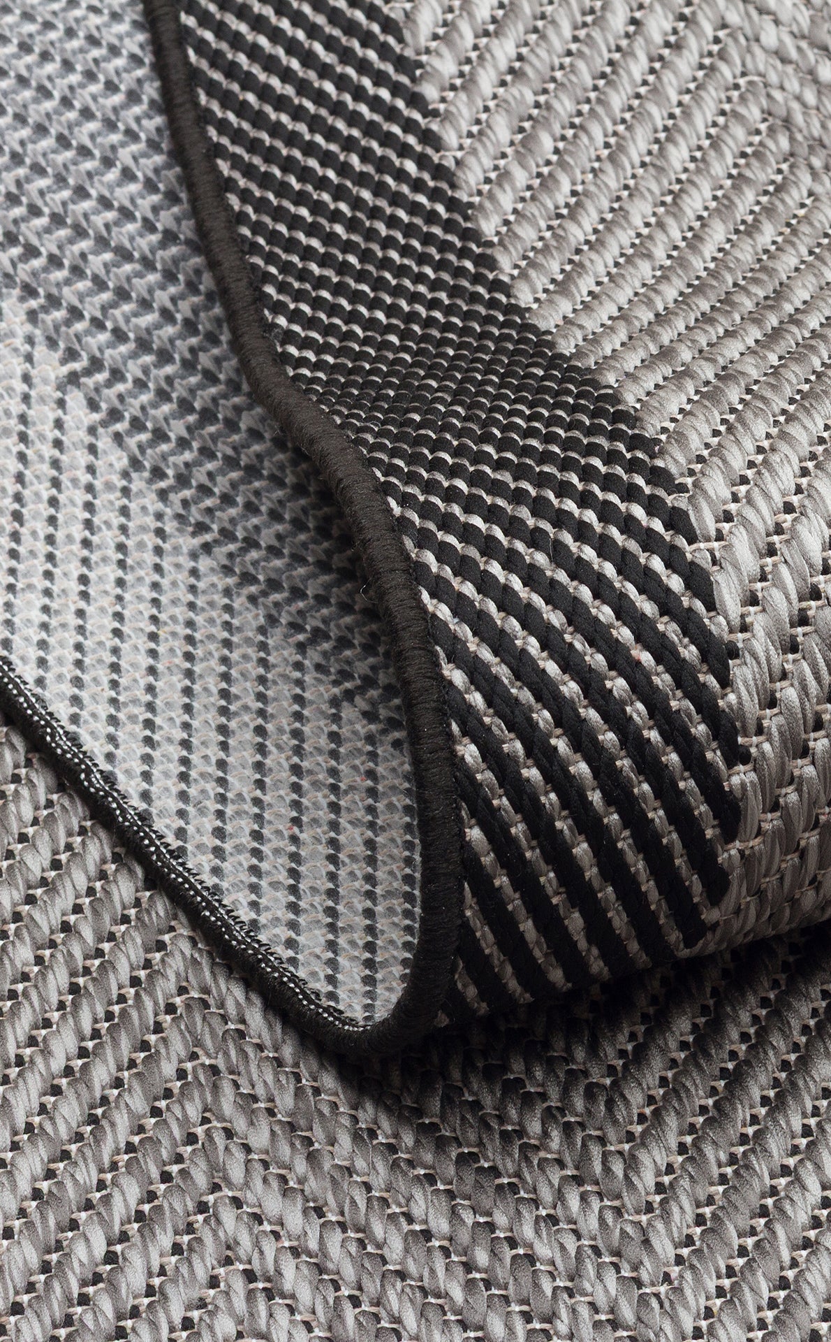 Close-up of Coal Texture - Kilim - MNO02, a woven fabric in gray and black tones with an anthracite kilim look; image shows its folded edge, revealing both sides of the detailed geometric pattern.