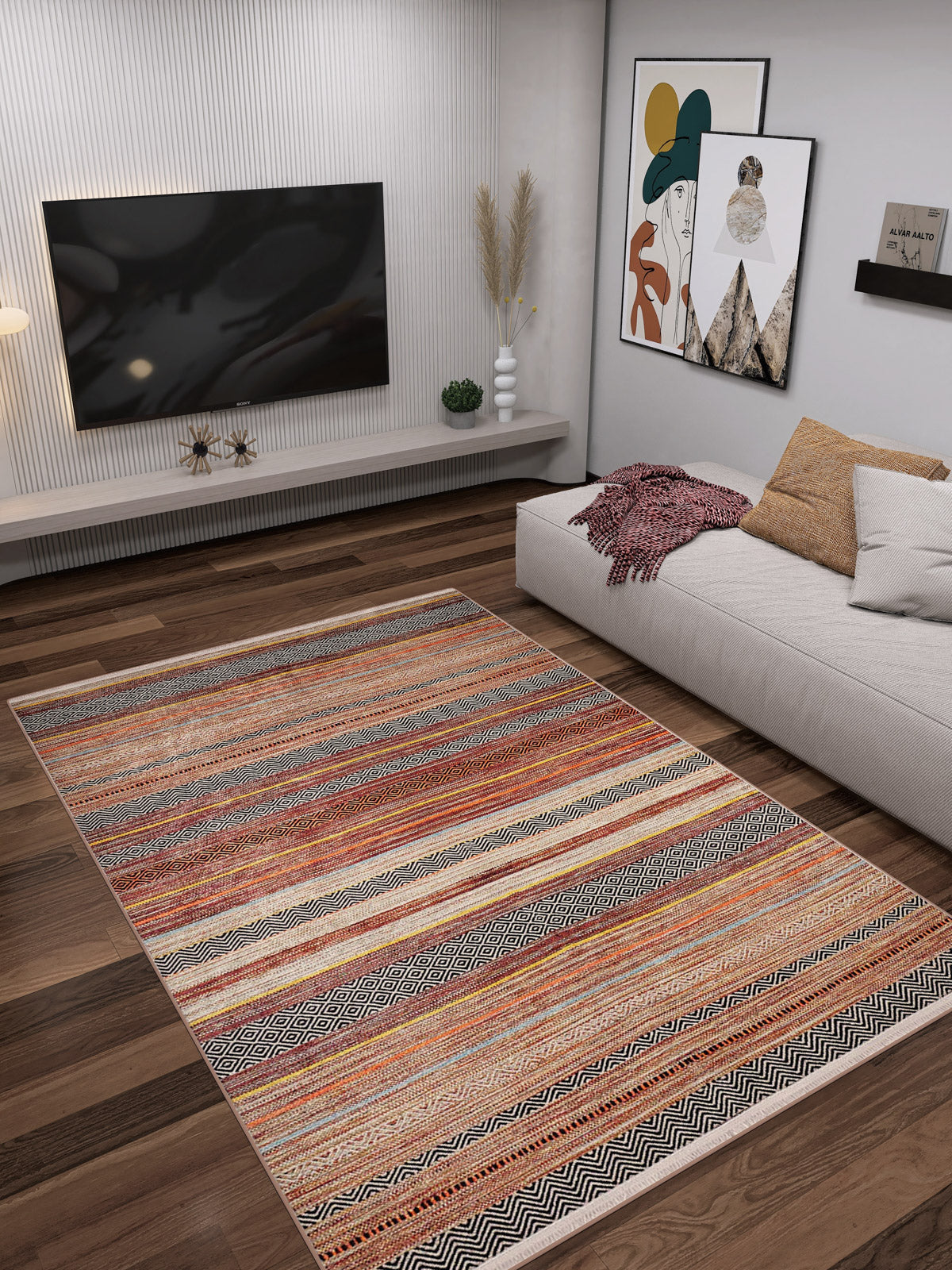 A modern living room features a wall-mounted TV, abstract art, a beige sectional with pillows and blanket, and the allergy-sensitive Coastal Breeze - Washable Rug - JR1549 in colorful stripes on dark wood floors. Plants decorate the low shelf.