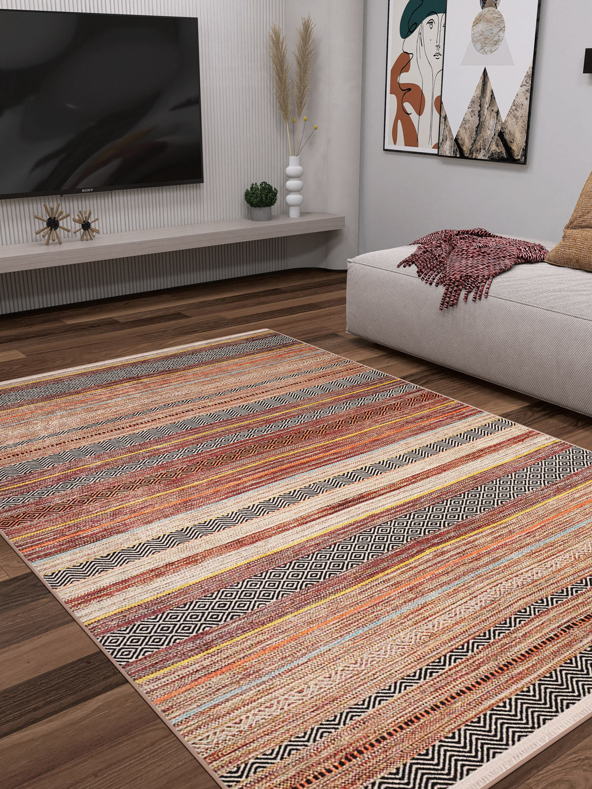 A modern living room featuring the Coastal Breeze - Washable Rug - JR1549 in bronze and brown earth tones, a low beige sofa with a red throw, wall-mounted TV, minimalist decor, framed art, and dried pampas grass—ideal for allergy-sensitive homes.