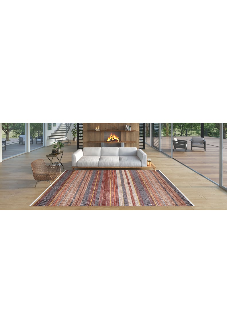 A modern living room features a white sofa, fireplace in a wooden wall, floor-to-ceiling windows, and the Coastal Breeze - Washable Rug - JR1549 (Custom Size)—perfect for high-traffic areas. A wicker chair sits on light wood floors with greenery outside.