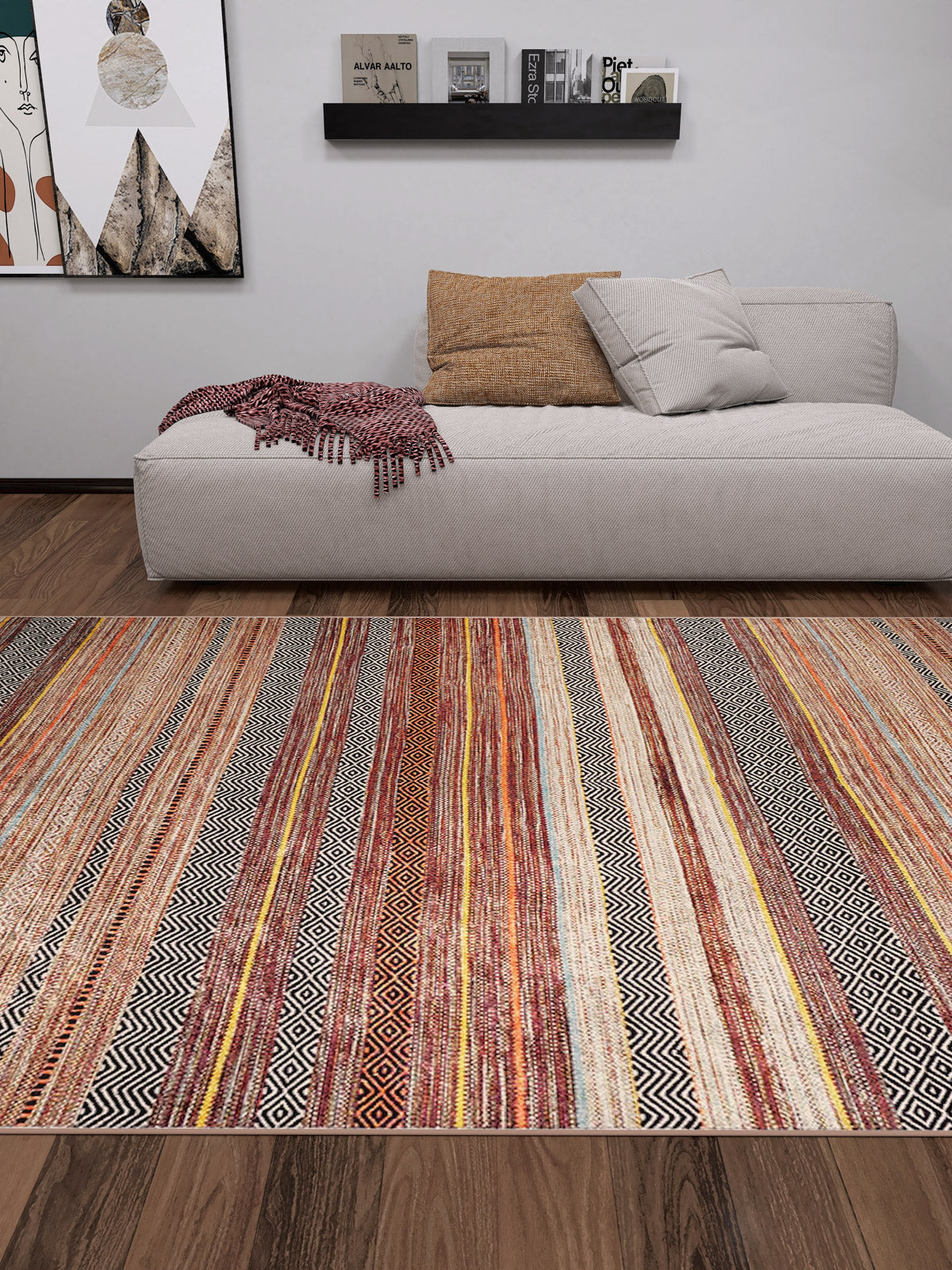 A modern living room features a beige sofa, tan pillow, red patterned throw, and the allergy-friendly Coastal Breeze - Washable Rug - JR1549 (Custom Size) in earthy tones—ideal for comfort and style. Wooden floor, wall art, and black shelf display books and decor.