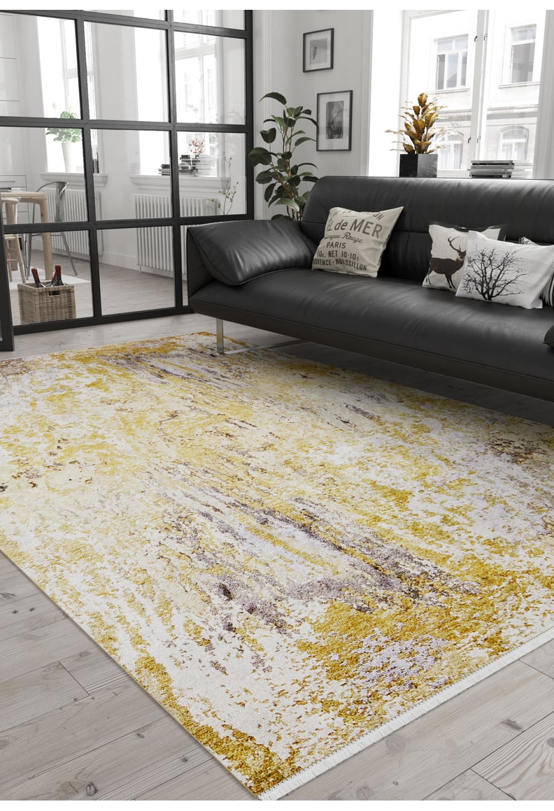 A modern living room features a black sofa, decorative cushions, potted plants, light wood floors, large windows with natural light, and the Color Splash - Washable Rug - JR146 (Custom Size) in yellow and white.