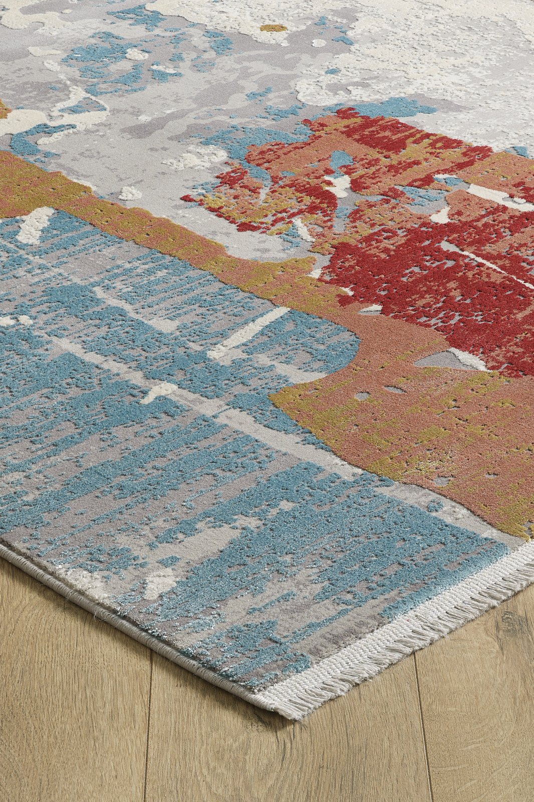 A close-up of the Colorful Chaos Premium Rug - M313P shows its abstract blue, red, gold, and white patterns, short fringe edging, and placement on a light wood floor.