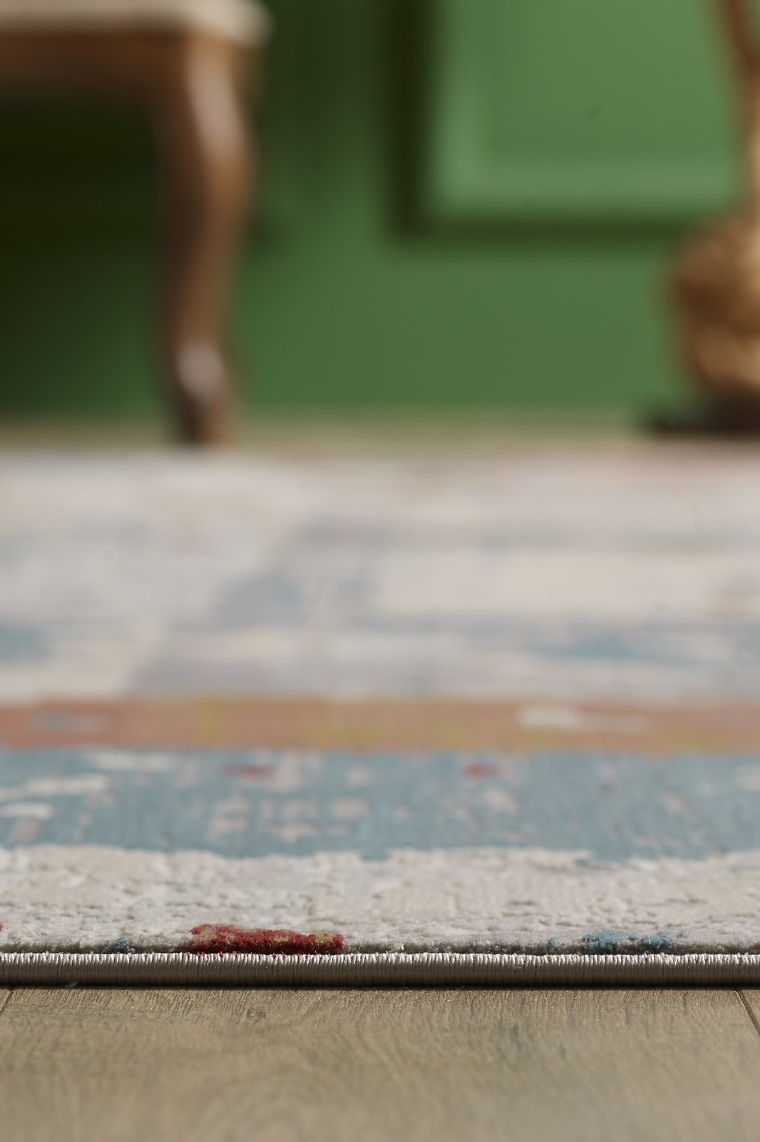 A close-up, low-angle view of the Colorful Chaos Premium Rug - M313P on a wooden floor, with a blurred green wall and chair in the background.