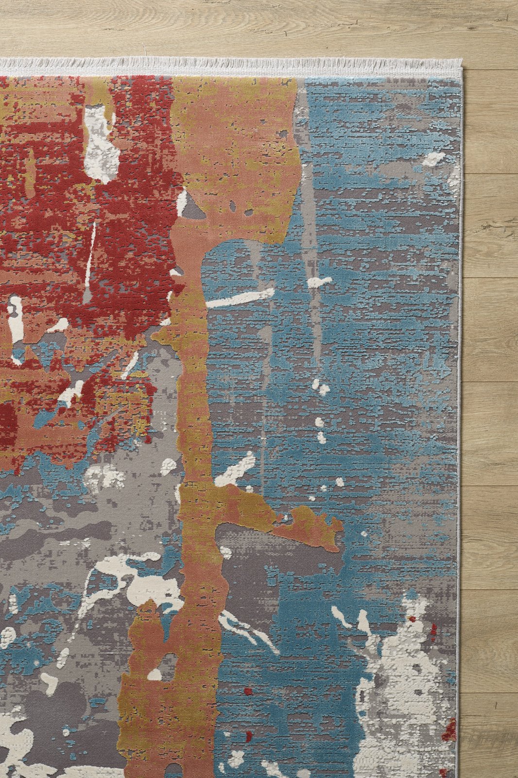 A corner of the Colorful Chaos Premium Rug - M313P on a wooden floor displays red, yellow, gray, white, and blue splashes in a painterly pattern with a white fringe along one edge.