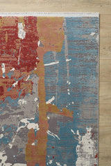 A corner of the Colorful Chaos Premium Rug - M313P on a wooden floor displays red, yellow, gray, white, and blue splashes in a painterly pattern with a white fringe along one edge.