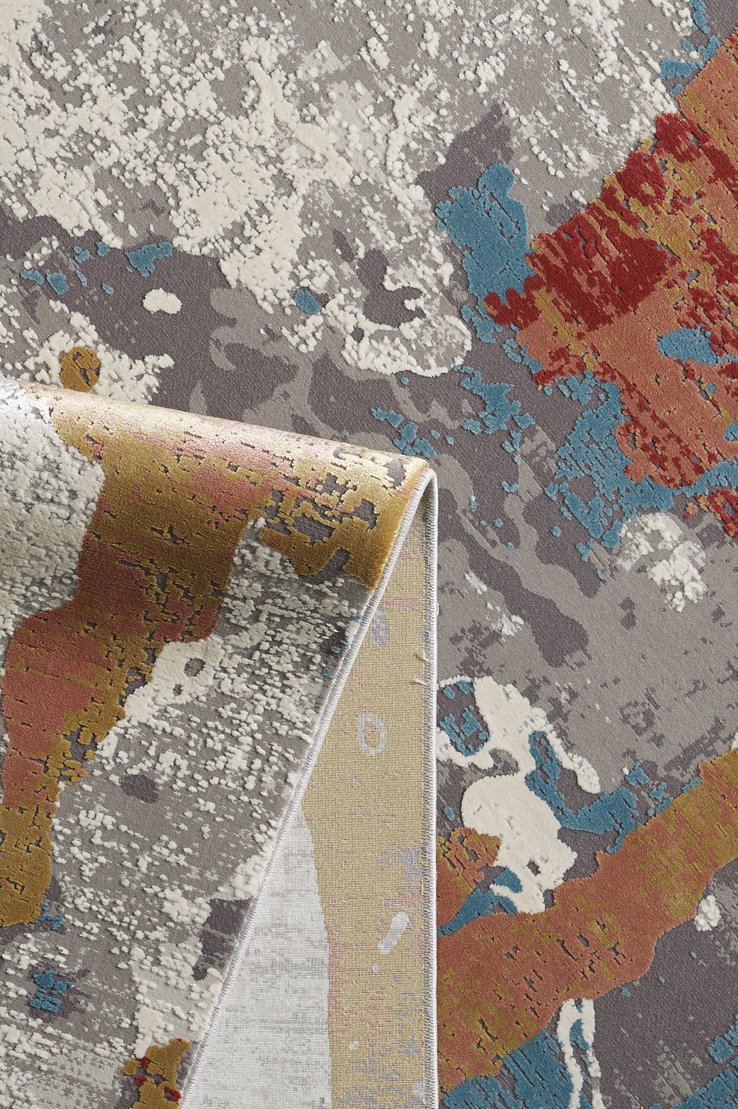 A close-up of the Colorful Chaos Premium Rug - M313P shows its modern multicolored pattern in gray, white, blue, red, and gold. One corner is folded over to highlight the rug’s machine-made underside and detailed edge stitching.