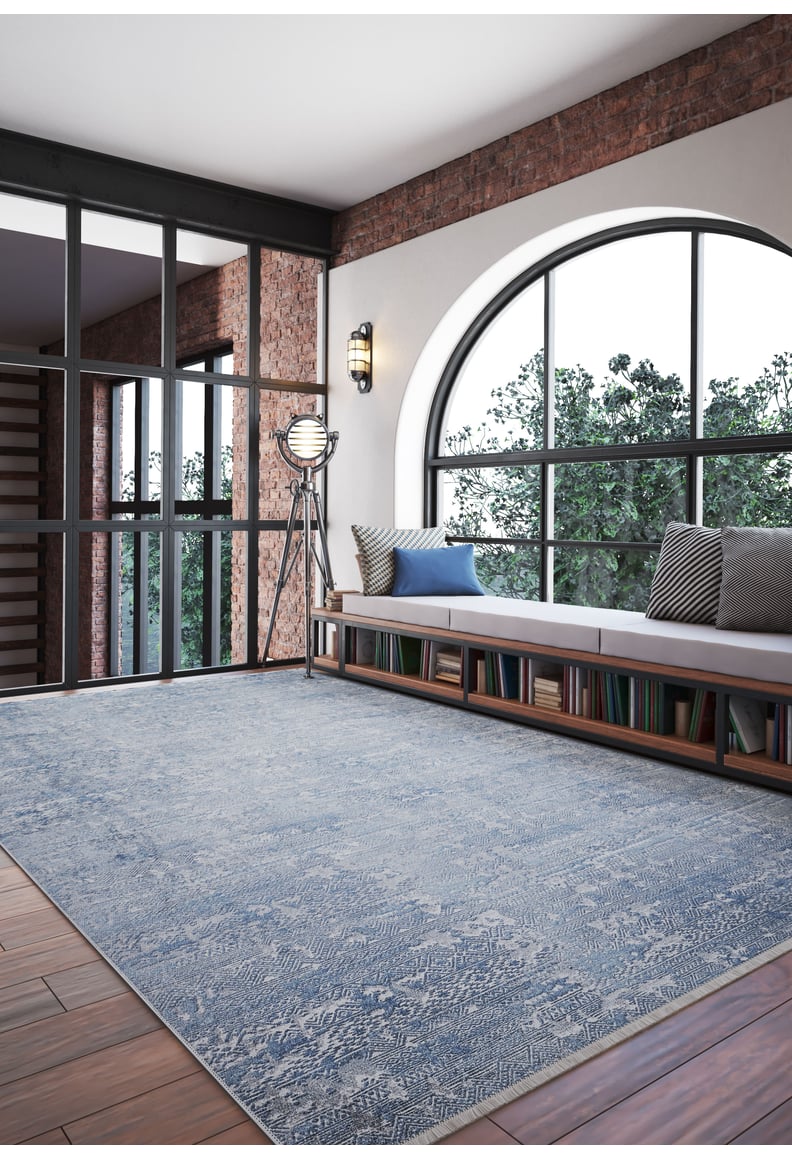 A modern room with a large arched window, brick walls, the Contemporary Serenity - Machine-Washable Rug JR1884 (Custom Size) in blue for high-traffic areas, a cushioned bench, bookshelves below, a vintage floor lamp, and trees visible outside.