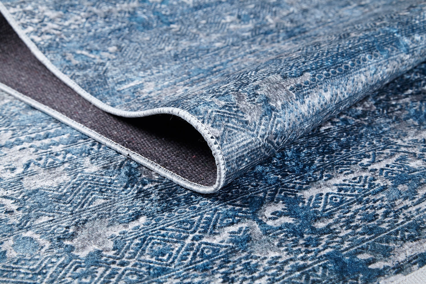 A close-up of the Contemporary Serenity - Machine-Washable Rug JR1884 (Custom Size) in blue and white geometric and floral patterns, partially folded to show the stitched edge—ideal for high-traffic areas.