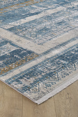 A close-up of the Cool Nomadica Premium Rug - M366B, a machine-made rug with blue, beige, and gold tones in geometric and floral patterns, fringed edges, and a subtle distressed finish on a light wood floor.