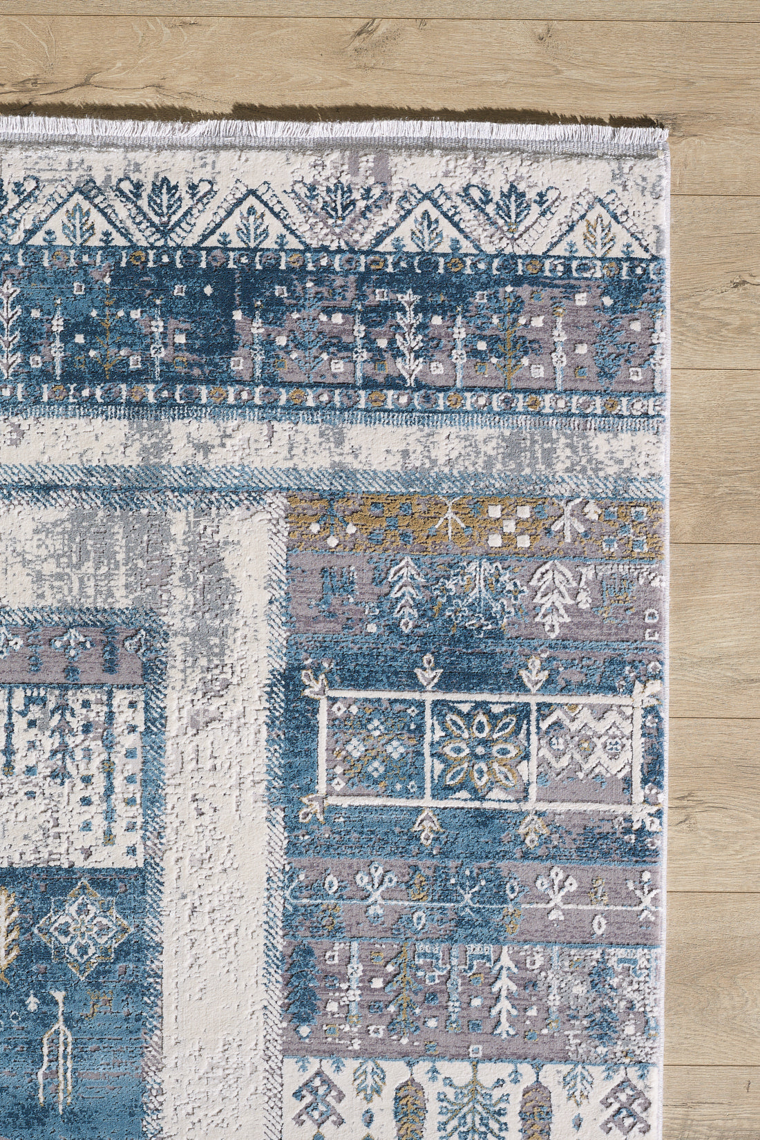 The Cool Nomadica Premium Rug - M366B features a decorative distressed finish with intricate blue, white, and beige geometric and floral patterns, symmetrical designs, and a textured look on a light wooden floor.