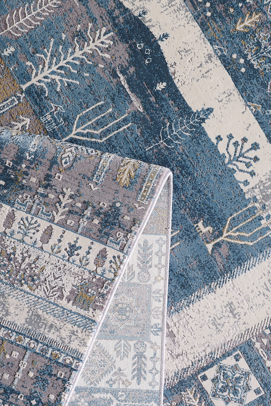 A close-up of the Cool Nomadica Premium Rug - M366B shows blue, beige, and grey geometric and botanical patterns. A folded corner displays a lighter underside design.