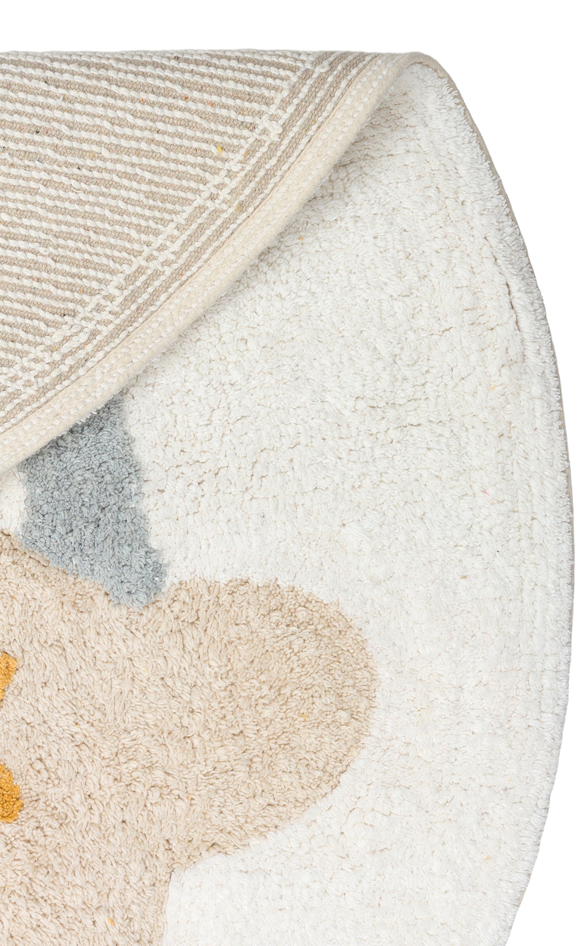 A close-up of the Cozy Puzzle - Washable Shaggy - CBN07 shows its round, mostly white surface with beige, gray, and yellow abstract shapes. Ideal for kids’ rooms, this rug is washable and features a striped underside with a folded corner.
