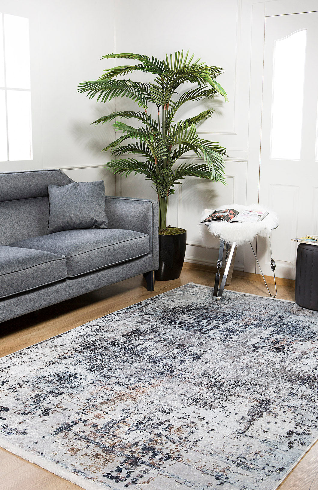 A modern living room featuring a gray sofa, the Creative Currents - Washable Rug - JR835 (Custom Size), a tall potted plant, and a side table with a magazine, all set against white walls with windows and a wooden floor.