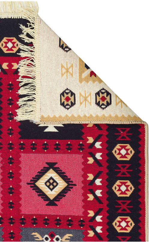 Load image 1 in gallery view. The Crimson Form - Washable Reversible Kilims - AR28 features tribal geometric patterns in red, black, cream, and yellow. A folded corner reveals its lighter underside with a similar design and fringe, highlighting its reversible style.