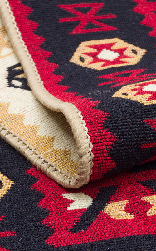 Load image 1 in gallery view. A close-up of the Crimson Form - Washable Reversible Kilims - AR28 showcases red, black, beige, and white tribal geometric patterns with a folded edge displaying detailed stitching and texture on this reversible rug.