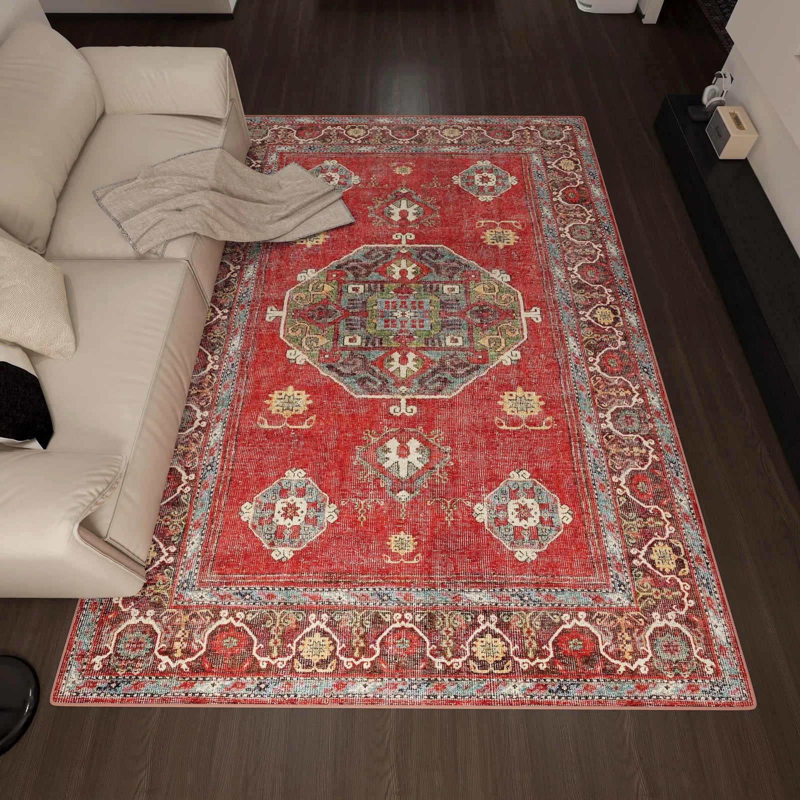 The Crimson Palace - Washable Rug - JRN1041 (Custom Size) with intricate red patterns lies on a dark wood floor next to a light sofa with white and beige cushions and a beige throw, complementing the room’s modern decor.