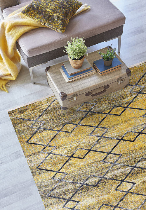Load image 1 in gallery view. A modern living room featuring the Crossed Connections - Washable Rug - JR1181 in yellow-gold, perfect for high-traffic areas, paired with a vintage suitcase coffee table, books, potted plants, a beige bench, and a yellow throw blanket.