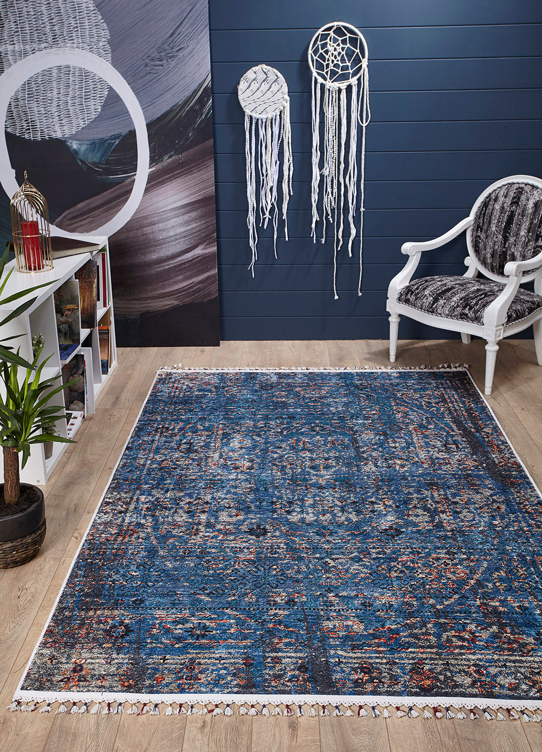A room with a Desert Diamond - Washable Rug - JR5071 (Custom Size) in blue pattern, a white chair and cushion, bookshelf, potted plant, two large white dreamcatchers on a dark blue wall, and light wood floors ideal for high-traffic areas.