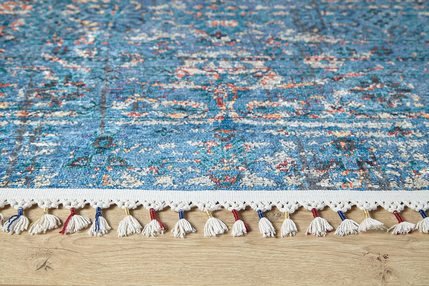 A close-up of the Desert Diamond - Washable Rug - JR5071 (Custom Size) in blue, with multicolored tassels and intricate blue, white, and red designs on a light wood floor—perfect for high-traffic areas.
