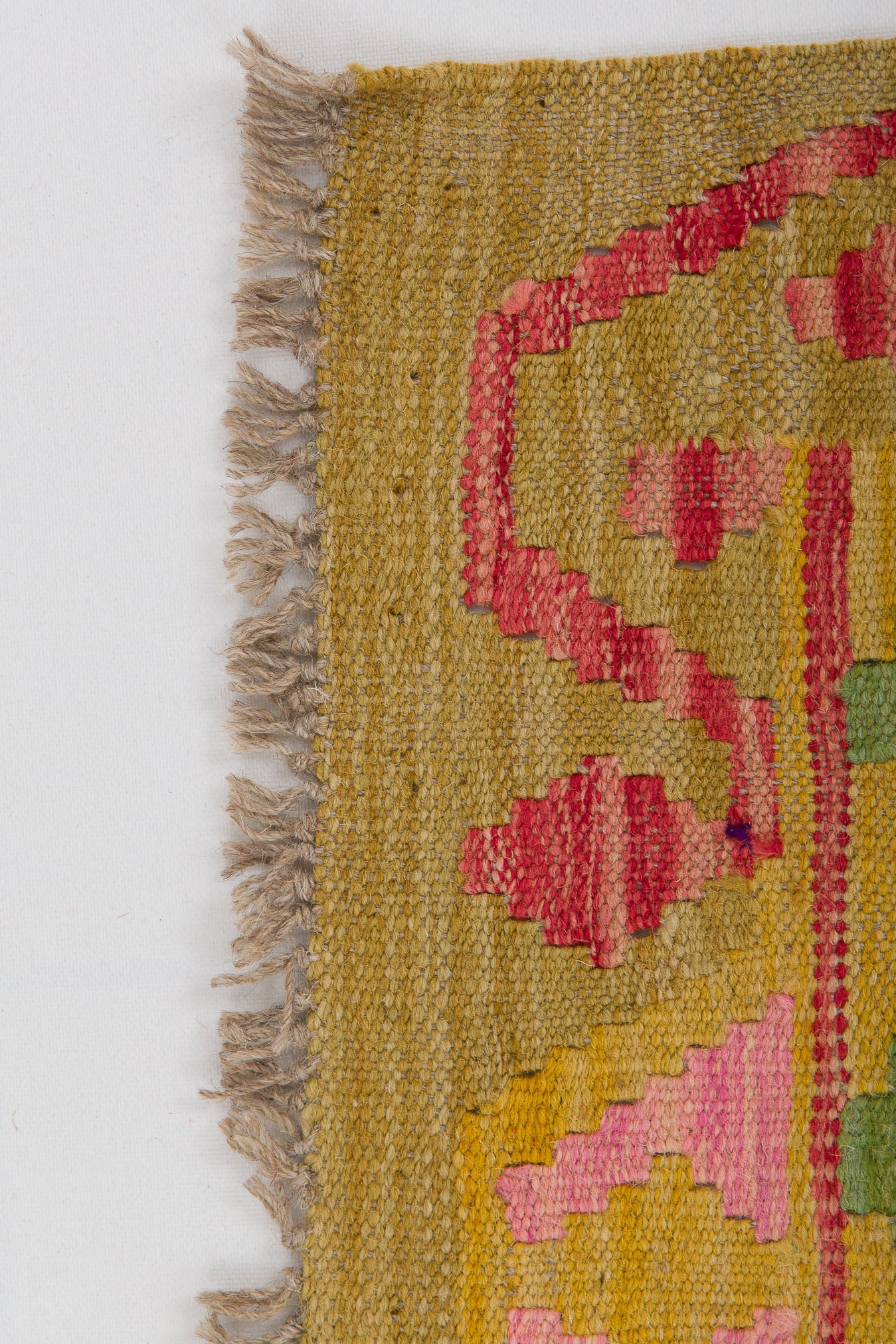 A close-up of the Desert Mirage - Handwoven Kilim reveals a corner with beige fringe and a geometric pattern in pink, red, yellow, and green on a muted tan background. The flatweave rug’s edge looks slightly worn.