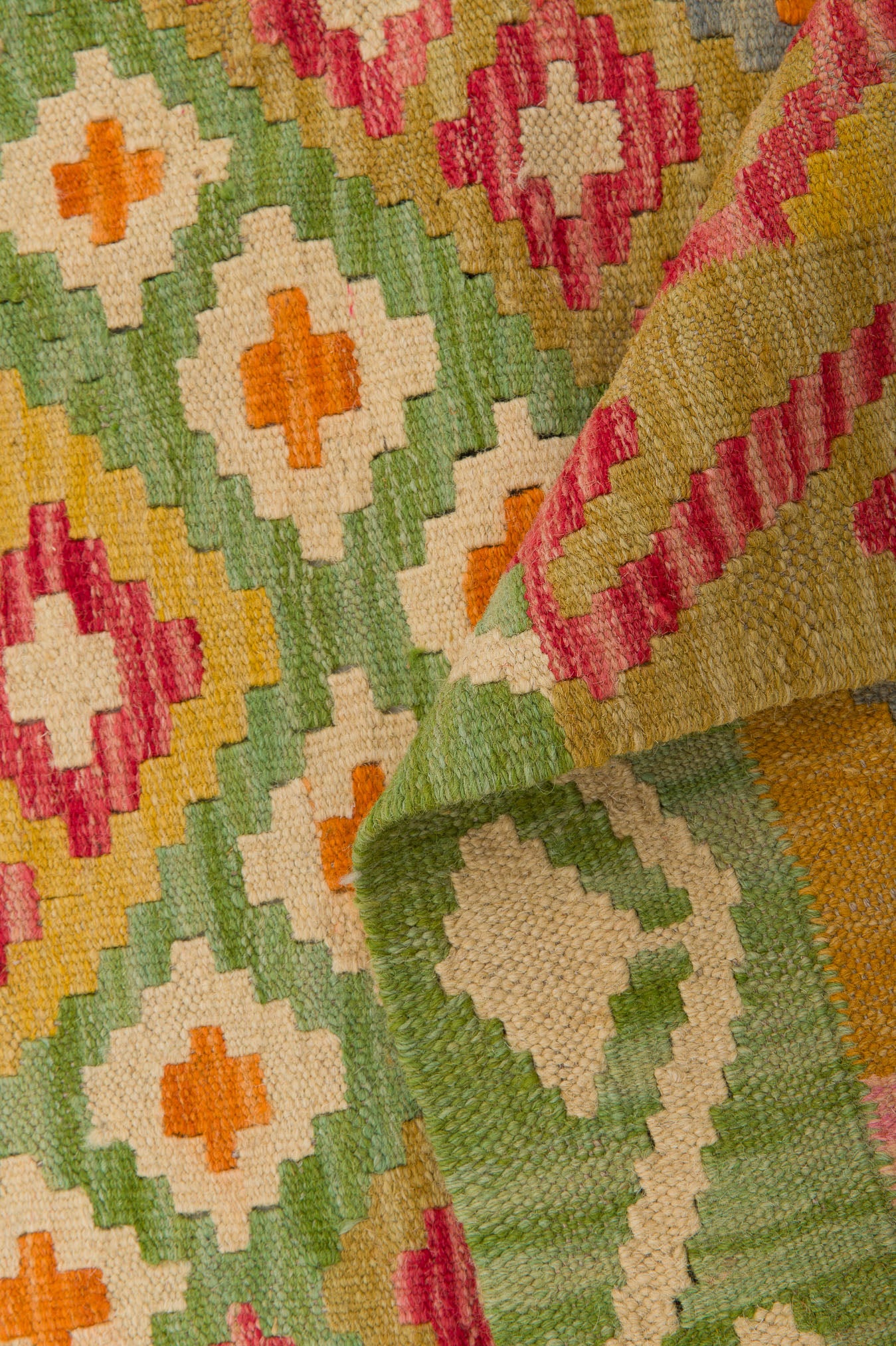A close-up of the Desert Mirage - Handwoven Kilim shows its colorful geometric patterns in green, orange, red, yellow, and cream. A folded corner reveals the textured weave and intricate flatweave design of this beautiful rug.