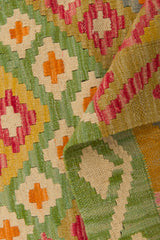 A close-up of the Desert Mirage - Handwoven Kilim shows its colorful geometric patterns in green, orange, red, yellow, and cream. A folded corner reveals the textured weave and intricate flatweave design of this beautiful rug.