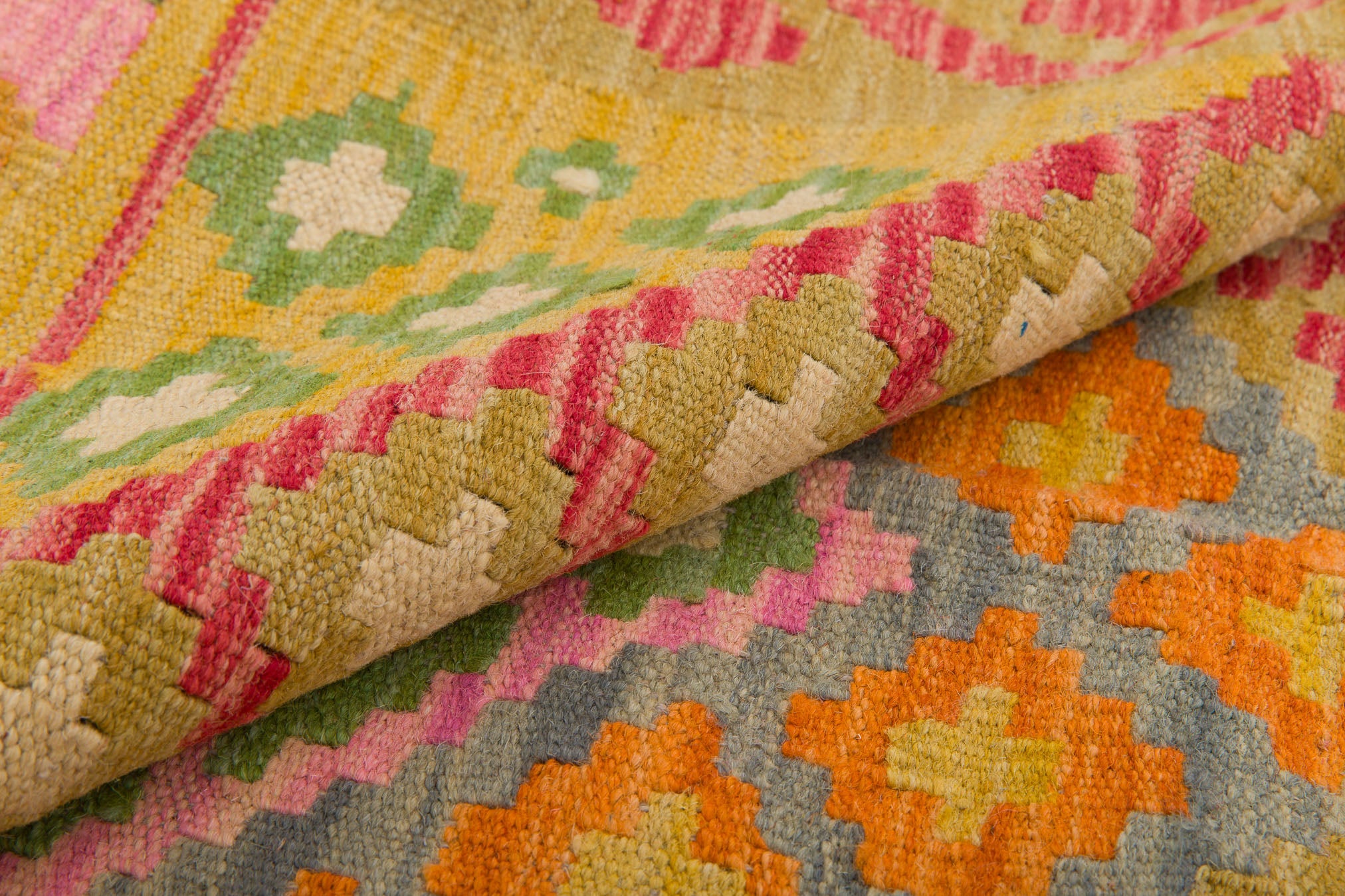 Close-up of the Desert Mirage - Handwoven Kilim, showcasing vibrant geometric patterns in yellow, green, white, pink, orange, and blue. A folded corner reveals the intricate flatweave design on both sides of this colorful rug.