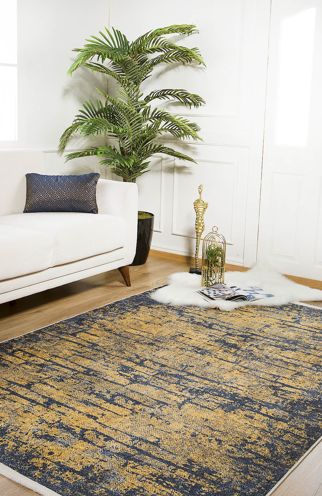 A modern living room with a white sofa, patterned cushion, large potted plant, Diagonal Dash - Washable Rug - JR1310 (Custom Size) in abstract blue and gold, white fur rug, decorative birdcage, golden statue, and magazines on a wooden floor.