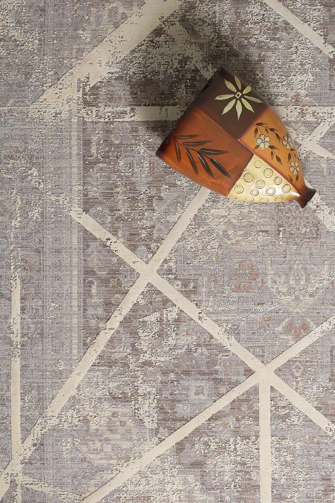 The Dominant Lineage Modern Rug - Bright - M633M features intersecting cream lines on gray for modern elegance, accented by a decorative ceramic vase with floral and leaf motifs in the top right corner.