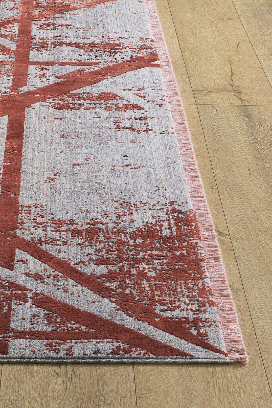 A corner of the Dominant Lineage Modern Rug - Coral - HRD003 shows abstract red and pink patterns on a light gray background with a pink border and clean edges, placed on a wooden floor for an industrial chic look.