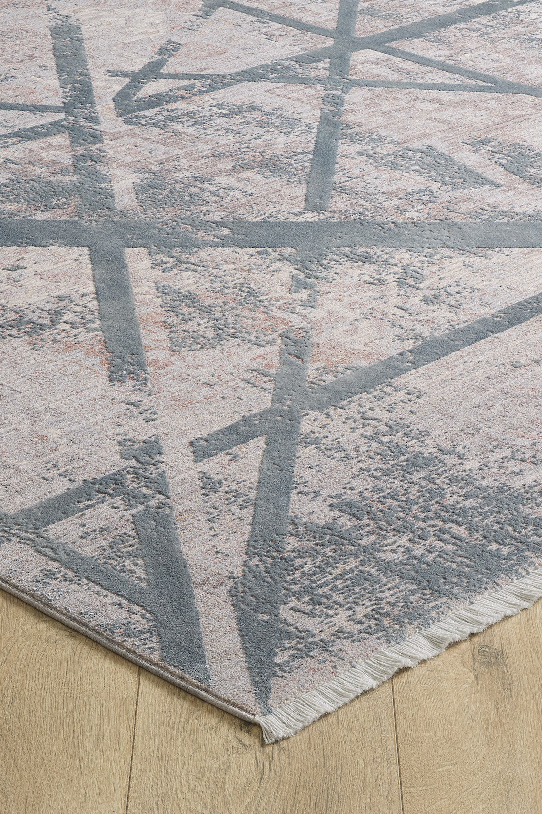 A close-up of the Dominant Lineage Modern Rug - Grey - HRD006, showcasing its geometric gray and light pink pattern with intersecting lines and a fringed edge, placed on a light wood floor.
