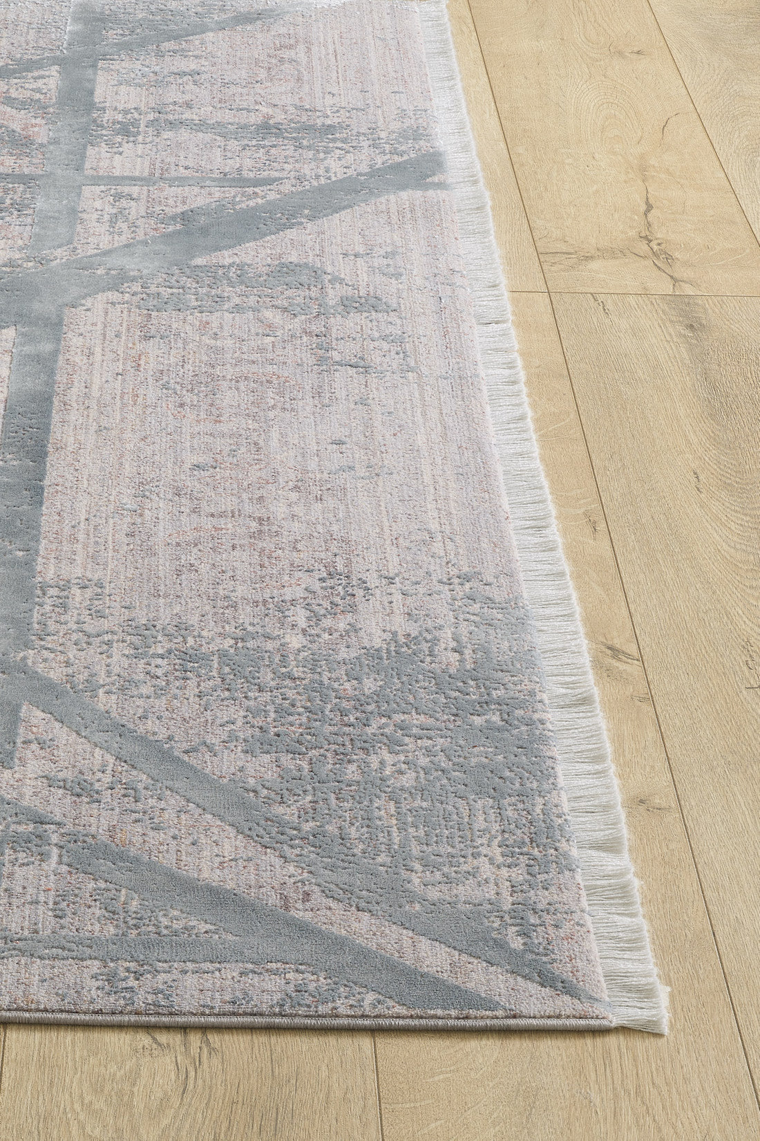 A close-up of the Dominant Lineage Modern Rug - Grey (HRD006) shows its light pink and gray abstract pattern and short white fringe, placed on a light wood floor.