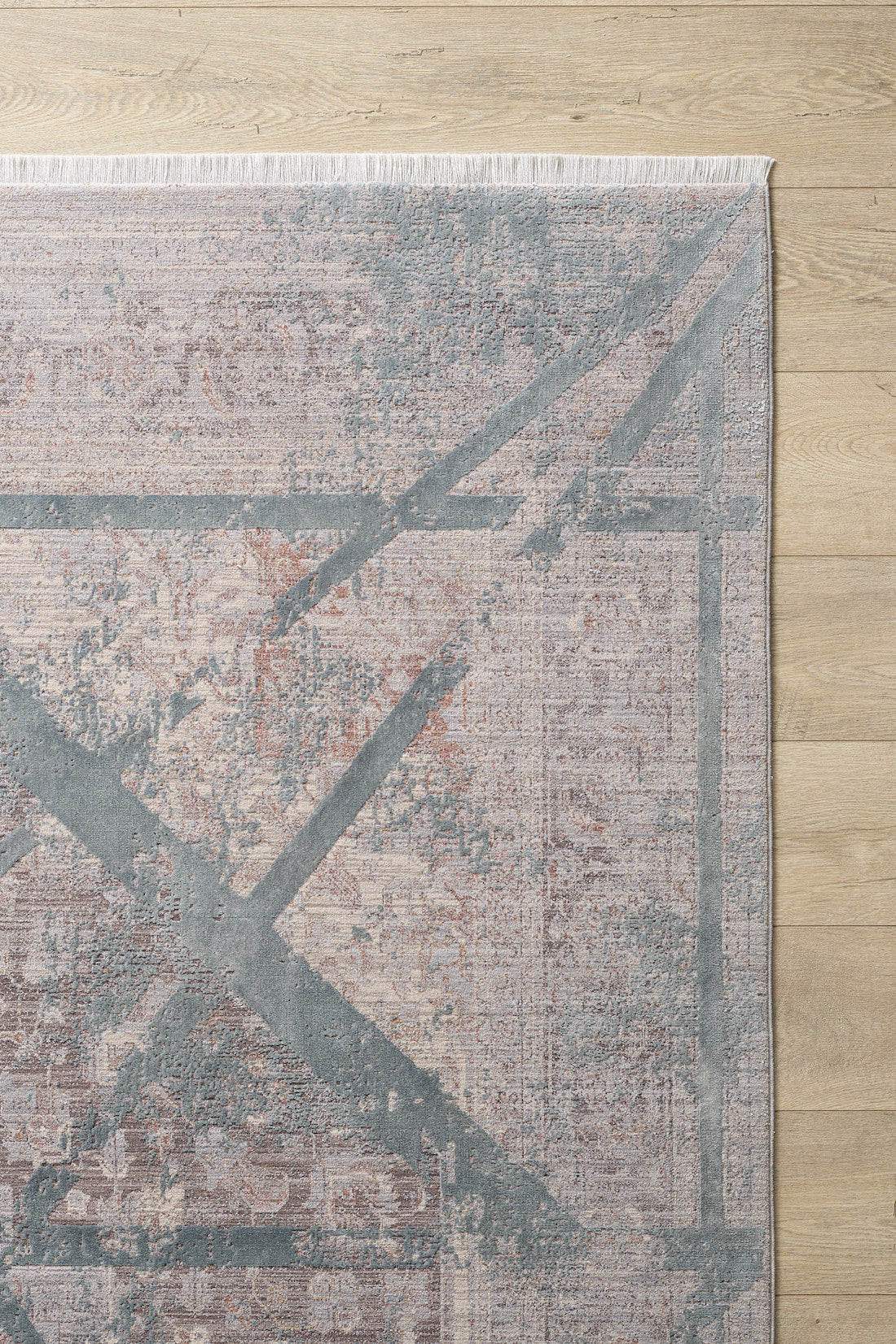 A close-up of the Dominant Lineage Modern Rug - Grey (HRD006), featuring an abstract geometric X pattern in muted gray and beige tones with top-edge fringe, displayed on a light wood floor.