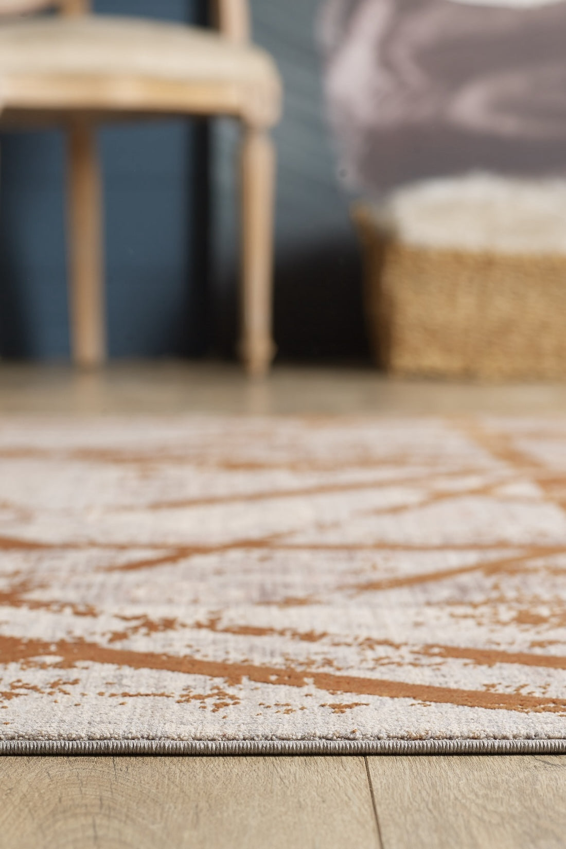 A close-up of the Dominant Lineage Modern Rug - Tangerine - HRD007 with beige and brown tones on a wooden floor. In the blurred background, a light wooden chair and woven basket sit against a dark wall.