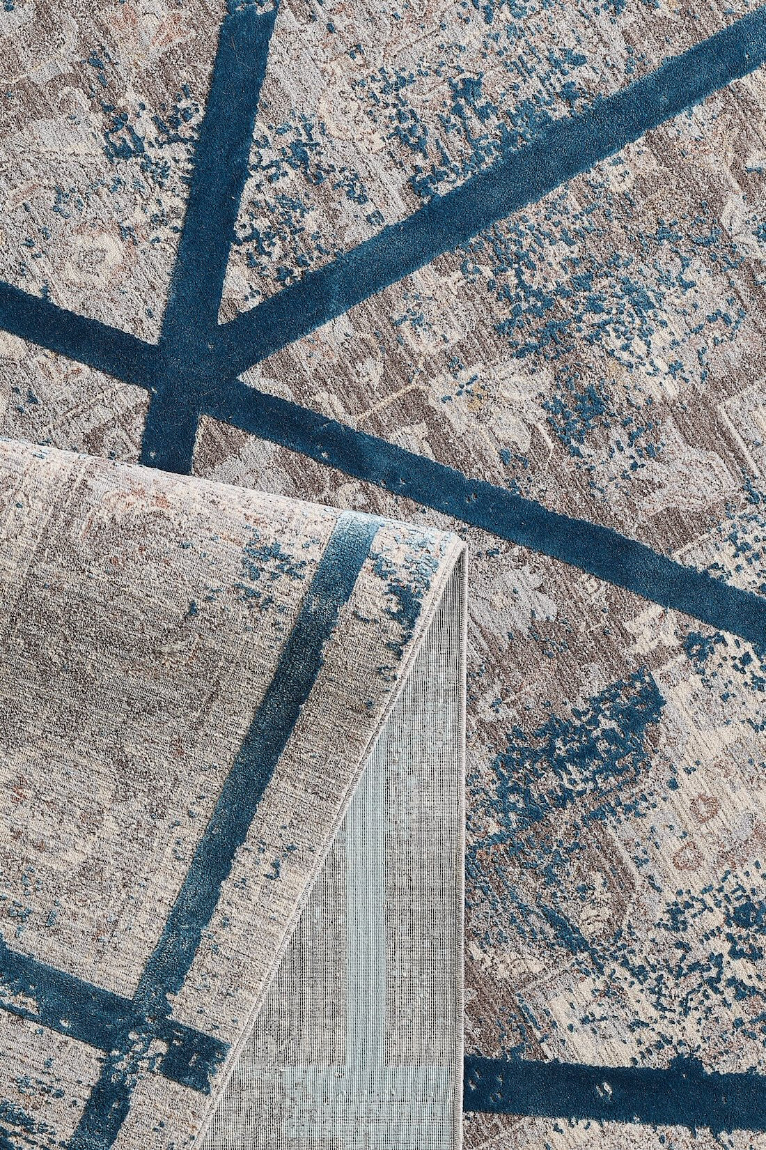 A close-up of the Dominant Lineage Modern Rug - Teal - HRD010 shows blue geometric lines and abstract beige and gray patterns, with one corner folded to reveal the machine-made underside.