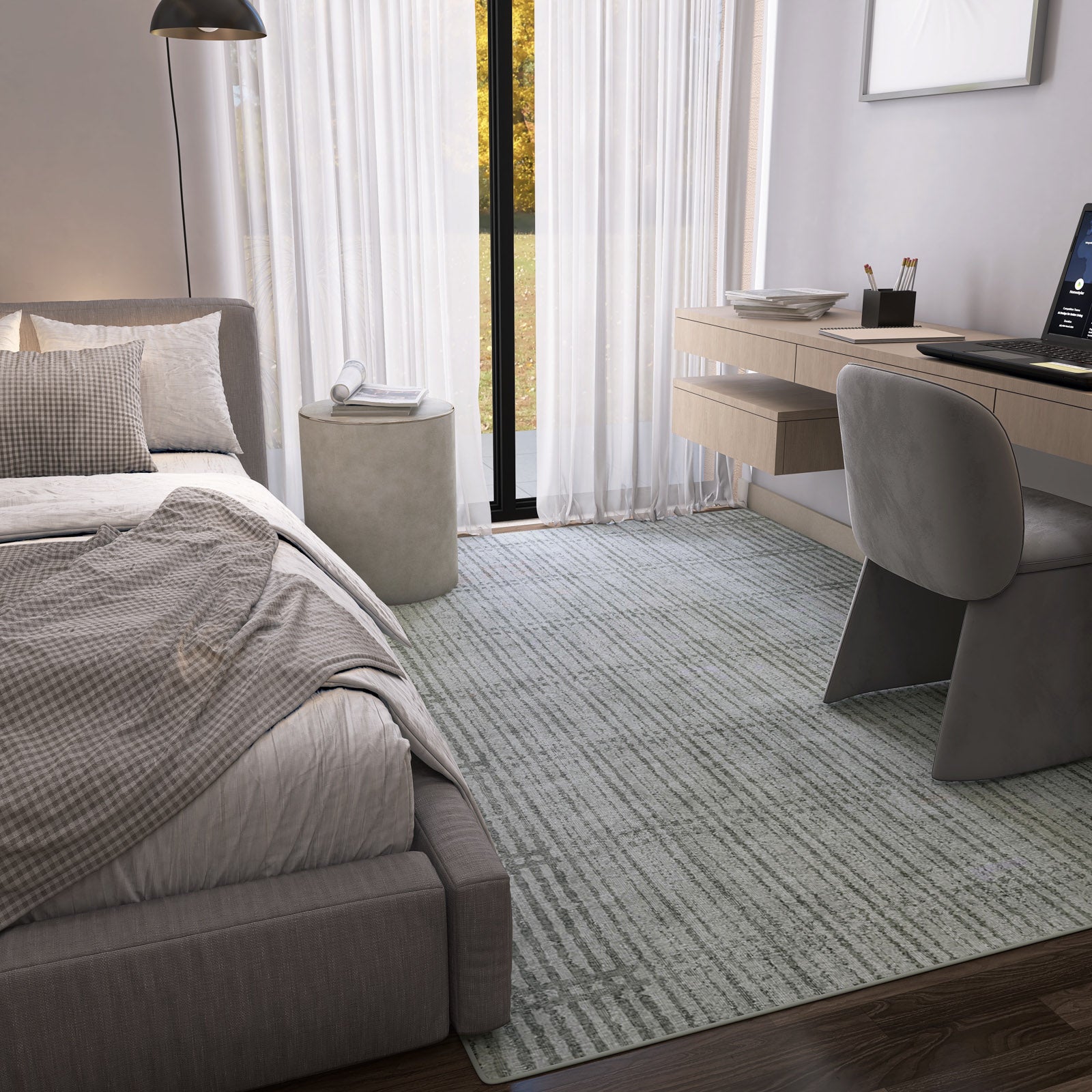 A modern bedroom with a neatly made bed, gray checkered blanket, Ornate Hues - Washable Rug - JRN38 for high-traffic areas, sheer white curtains on tall windows, a floating desk with computer and gray chair. Natural light brightens the space.