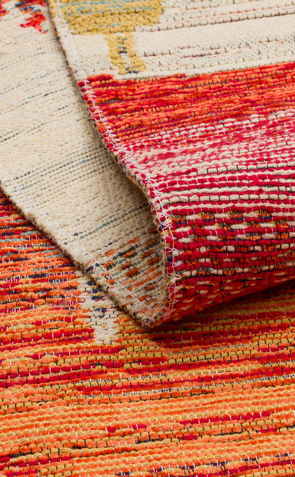 A close-up of the Dust Trace - Kilim - NMD12 rug with a folded corner, showcasing its vibrant red, orange, yellow, and cream hues in a textured, patterned modern design.