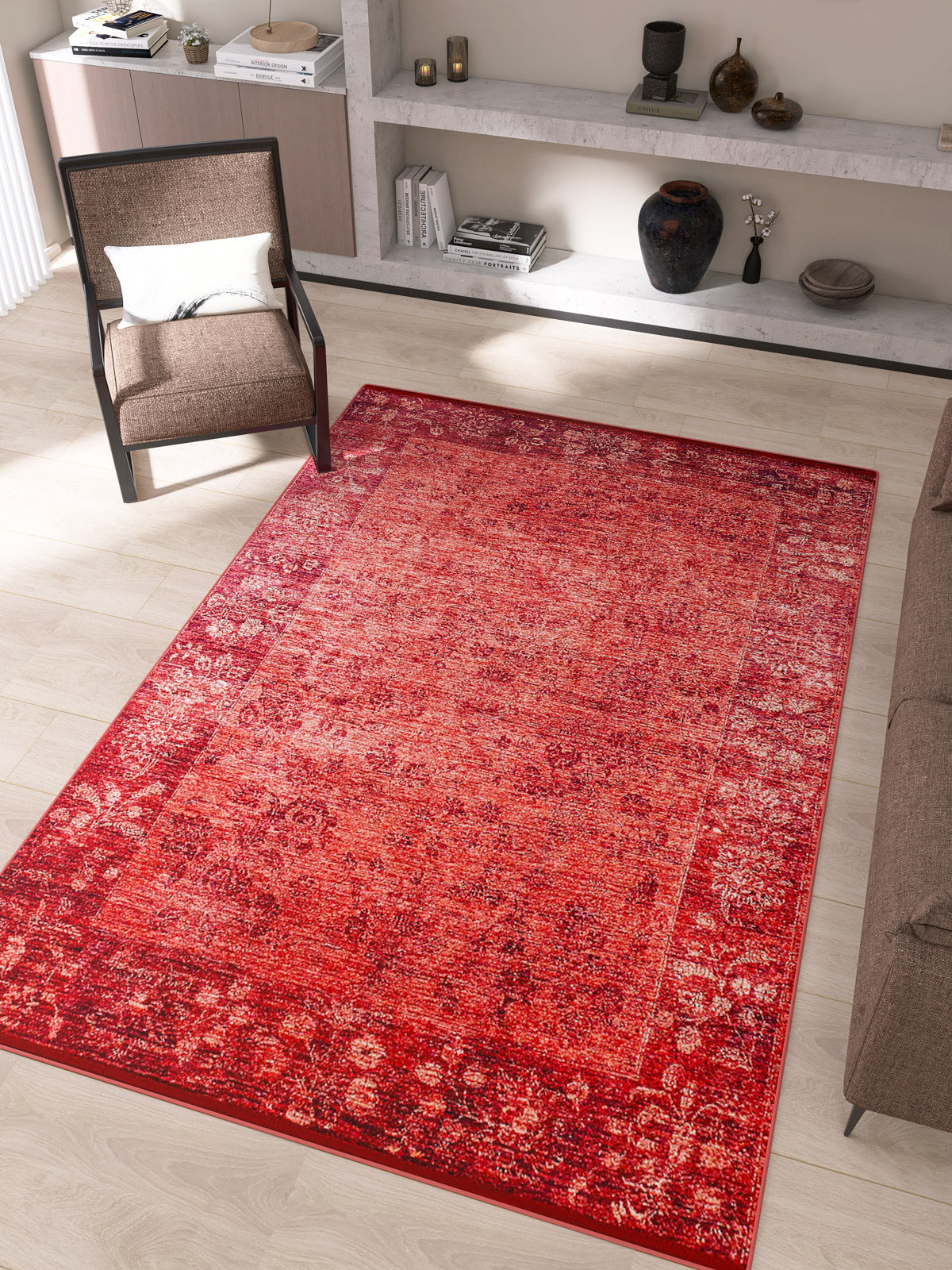 A living room featuring the Dynasty Dream - Washable Rug - JR160 in a red oriental pattern, a brown armchair with a white pillow, beige couch, and shelves with vases, books, and decor against light walls and a wooden floor.