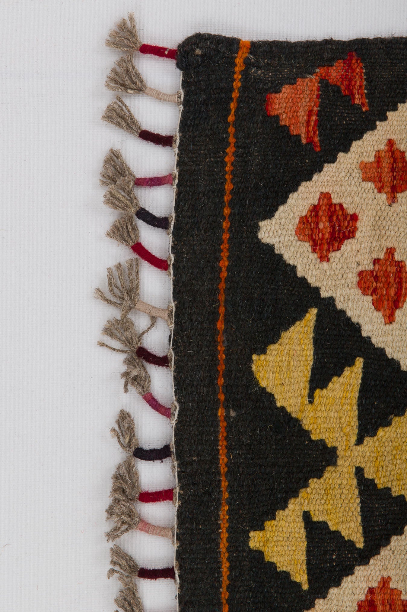 A close-up of the Earthly Lines - Handwoven Kilim shows geometric patterns in yellow, orange, and red on a black background, with knotted fringe along the left edge.