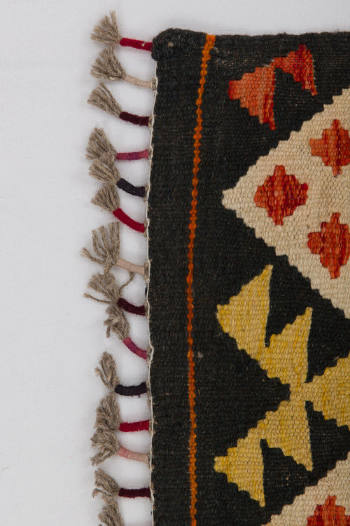 Load image 1 in gallery view. A close-up of the Earthly Lines - Handwoven Kilim shows geometric patterns in yellow, orange, and red on a black background, with knotted fringe along the left edge.